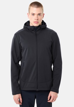 Junger Mann trägt eine schwarze Kapuzenjacke mit Reißverschluss und seitlichen Reißverschlusstaschen, steht vor einem schlichten Hintergrund.