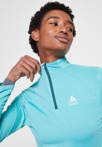 Turkoois kleurige rits-tot-top met lange mouwen, gemaakt van een glad materiaal. Het heeft een hoge kraag, blauwe rits en een subtiel logo op de borst.