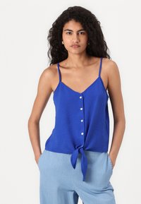 Haut sans manches bleu avec un devant noué, présentant un tissu texturé, un décolleté en V et six boutons blancs sur le devant.