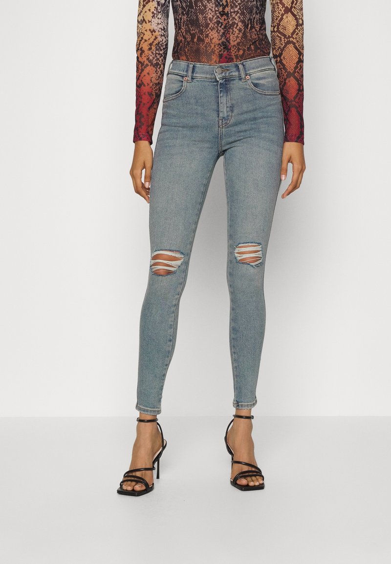 Lichtblauwe skinny jeans met versleten scheuren bij de knieën, met een hoge taille en subtiele vervaging. Gecombineerd met zwarte stringhakken.