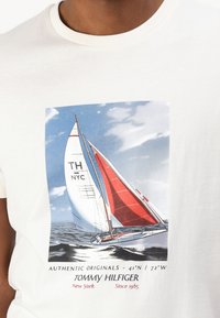 Camiseta de algodón blanca con un gráfico de un velero con una vela roja y un cielo azul, etiquetada con "TH NYC" y detalles de marca.