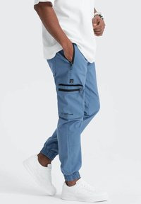 Pantalons cargo bleu clair avec poches zippées et poignets élastiques, associés à des baskets blanches. Le matériau semble durable et respirant.