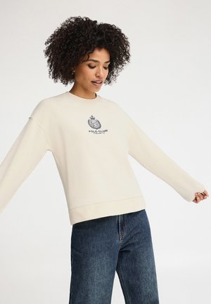 Donna con capelli ricci che indossa una felpa bianco sporco con il logo Polo Club e jeans blu, braccia leggermente aperte, su uno sfondo uniforme.