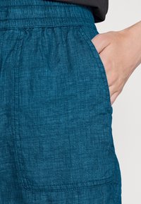 Pantalon en lin bleu sarcelle avec une ceinture élastique, doté d'une poche latérale et d'un tissu texturé. Des coutures visibles soulignent le design.