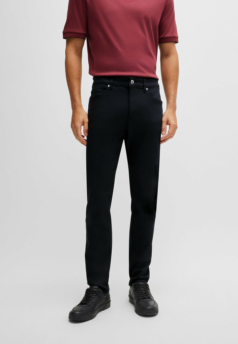 Pantalons noirs en mélange coton avec une coupe droite, cinq poches, bouton en métal et détail de logo, associés à une chemise bordeaux à manches courtes.
