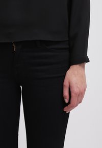 Svart långärmad blus med rundad nederkant, kombinerad med mörka jeans. Synlig dragkedja vid midjan; slät textur.