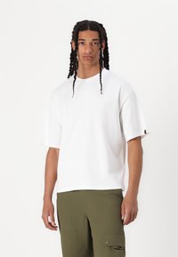 T-shirt en coton blanc à manches courtes, coupe décontractée, avec une petite étiquette logo sur la manche gauche. Associé à un pantalon cargo vert olive.