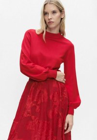 Czerwony dziany sweter z wysokim kołnierzem i długimi, obszernymi rękawami, połączony z czerwoną kwiatową koronkową spódnicą, tworząc efekt warstwowy.