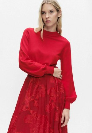 Röd stickad tröja med hög krage och långa, voluminösa ärmar, kombinerad med en röd blommig spetskjol som skapar en lager-på-lager-effekt.