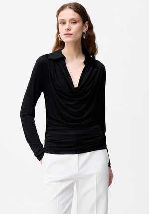 Kobieta ubrana w czarną, długorękawową bluzkę z kołnierzykiem i białe spodnie, stoi z rękami w kieszeniach na jednolitym tle.