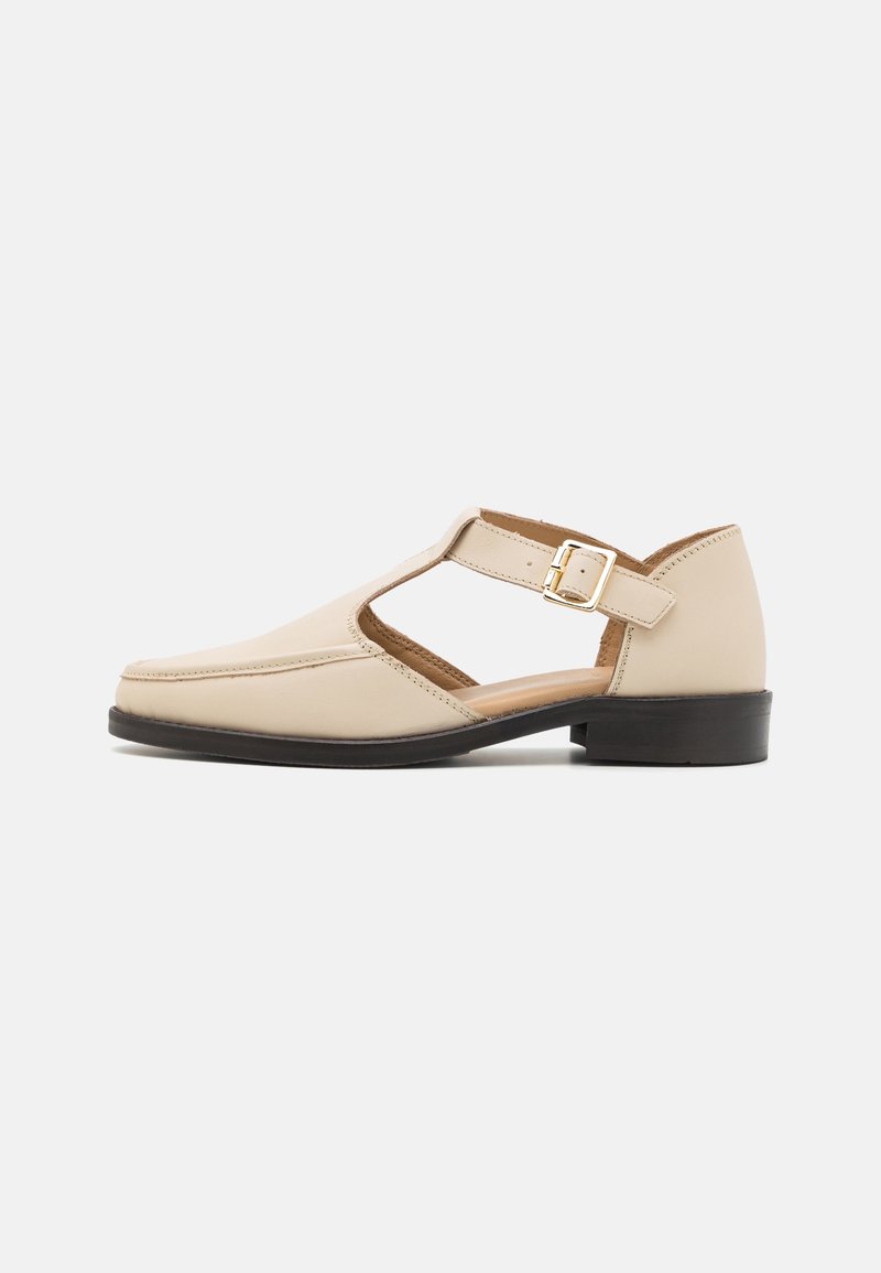 Chaussure en cuir beige avec un bout arrondi, un design ajouré et une bride de cheville ornée d'une boucle dorée. Talon bloc noir bas.