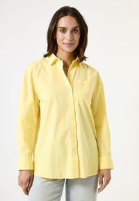 Camisa amarilla de botones con mangas largas, hecha de una tela ligera. Cuenta con un bolsillo en el pecho y un corte relajado, combinada con jeans de mezclilla azul claros.