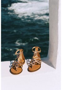 Sandalias de tiras en tono dorado con un diseño texturizado y suelas planas, que cuentan con lazos decorativos y correas para el tobillo, situadas en un fondo costero.