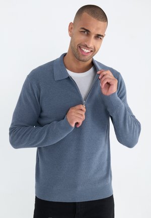 Maglione blu con zip, caratterizzato da una trama a coste, dotato di colletto piatto e maniche lunghe, indossato sopra una t-shirt bianca e abbinato a pantaloni scuri.