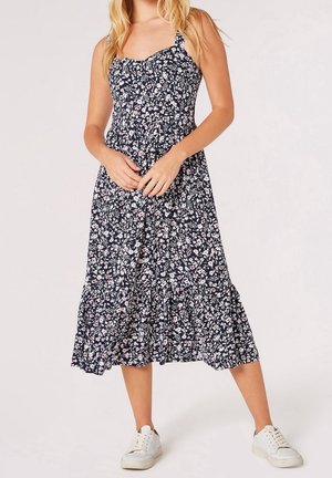 Vrouw draagt een marineblauwe midi-jurk met witte en roze bloemenprint, spaghettibandjes en witte sneakers, staand tegen een effen achtergrond.