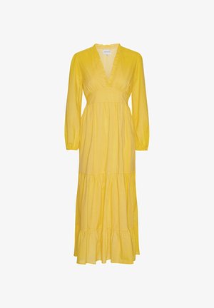 Femme portant une longue robe jaune à manches longues et col volanté, debout devant un fond beige uni.