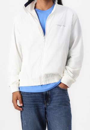 Personne portant une veste blanche zippée sur une chemise bleue et un jean bleu, une main dans la poche et l'autre détendue sur le côté.