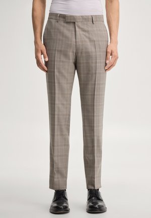Man die beige geruite nette broek en zwarte leren schoenen draagt, staand voor een effen lichte achtergrond.