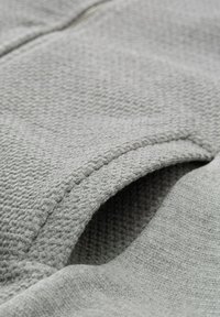 Grijze stof met een gestructureerd, wafelachtig patroon. Bevat een glad, vlak gedeelte en een naad langs de rand, wat de stiksels van het kledingstuk accentueert.