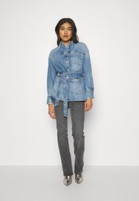 Denim srajca-jakna v svetlo modri barvi z zapenjanjem na pasu, ki ima ovratnik, sprednje žepe in gumbno zapiranje. Nosi se z sivo jeansom in metalnimi čevlji.
