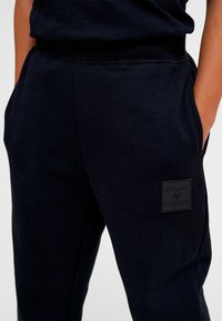 Sorte joggingbukser lavet af en bomuldsblanding, med sidelommer og et tekstureret patch-logo på låret. Elastisk taljebånd for komfort.