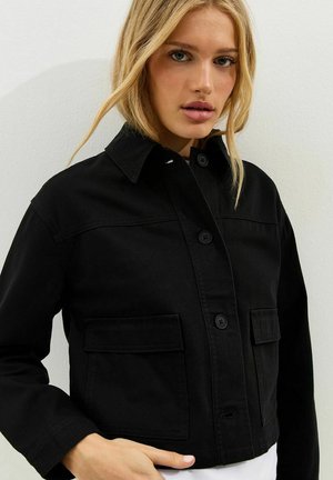 Blondynka w czarnej kurtce na guziki z dużymi przednimi kieszeniami, stojąca na tle jednolitej jasnej ściany.