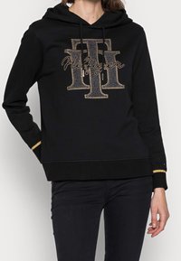 Sudadera negra con un logo de strass en la parte delantera, capucha con cordones y puños con detalles en dorado. Fabricada con un tejido suave y texturizado.