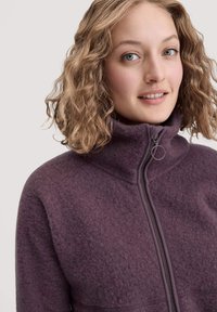 Nuori nainen, jolla on kihara vaaleanpunainen hius, pukeutunut vetoketjulliseen purppuraan fleece-takkiin, hymyilee lempeästi yksinkertaista taustaa vasten.