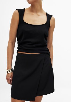 Zwarte mouwloze top met een vierkante halslijn, gecombineerd met een zwarte wikkelrok met een asymmetrische zoom en een gladde textuur. Zilveren armband.