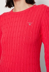 Woman wearing a bright red cable-knit sweater with a round neckline and a small GANT logo on the chest.