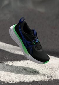 Chaussure de sport noire avec une tige texturée, des accents verts et bleus, une semelle intermédiaire blanche et un design épuré, suspendue au-dessus d'une surface.