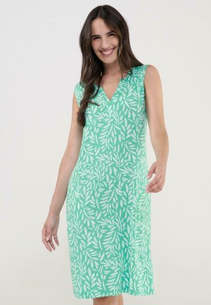 Mujer sonriente con el cabello largo y oscuro, vestida con un vestido verde sin mangas con patrón de hojas blancas sobre un fondo liso.