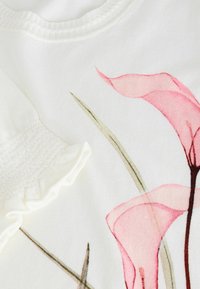 Witte stoffen top met een zachte, roze bloemenprint en groene bladeren; korte pofmouwen met een gerimpelde rand en een geribbelde halslijn.