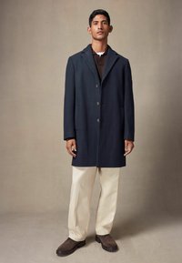 Abrigo azul marino con cuello muesca, cinco botones y bolsillos inclinados, superpuesto a una blusa marrón, combinado con pantalones crema y zapatos marrones.