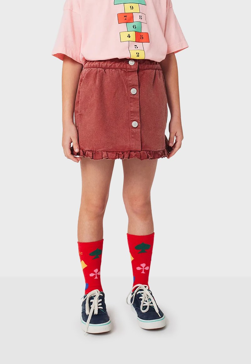 Jupe en denim rouge avec un ourlet volanté, dotée d'une fermeture à boutons sur le devant. Associée à des chaussettes colorées et des baskets bleu marine. Chemise rose à manches courtes.