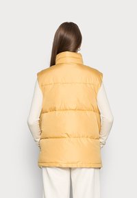 Gelbe Steppweste mit hohem Kragen, gestepptem Design und glatter Textur. Getragen über einem weißen, gemusterten Langarmhemd.