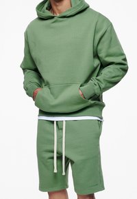 Sudadera verde con bolsillo frontal y pantalones cortos a juego. Fabricada con un tejido suave, presenta un pequeño logo, con un corte relajado y cordones.