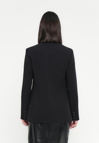 Donna con capelli scuri e lisci, che indossa un blazer aderente nero e una gonna di pelle nera, vista di spalle su uno sfondo bianco.