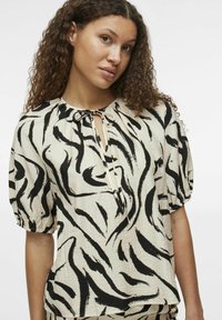 Blusa de manga abullonada en un crema claro, con un patrón de pinceladas negras. Escote en forma de ojal con detalle de lazo. Tejido ligero y texturizado.