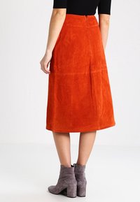 Oranssi mokkanahkainen midi-hame, jossa on sileä pinta, sivutaskut ja vetoketju takana. Yhdistetty harmaisiin nilkkureihin.