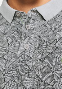 Polo gris avec un motif de feuilles noires, doté d'un col et de trois boutons sur le devant. Tissu texturé avec des détails subtils.