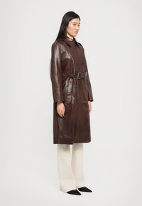 Femme aux longs cheveux noirs portant un trench-coat en cuir marron foncé ceinturé, un pantalon crème et des chaussures noires à bout pointu sur un fond uni.