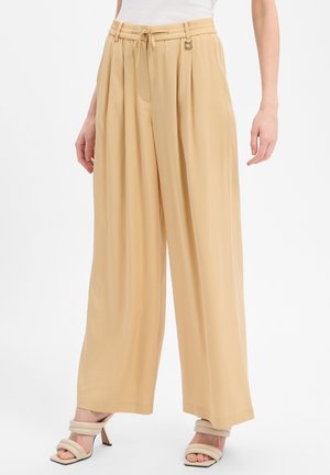 Beige wijde broek met treklint in de taille en plooien, gedragen met beige open-teen sandalen met hak en een wit topje.