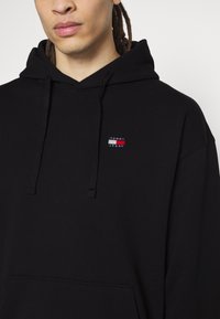 Schwarzer Hoodie mit einer vorderen Tasche, Kapuze mit Kordelzug und einem kleinen rechteckigen Logo-Patch in Rot, Weiß und Blau auf der Brust.