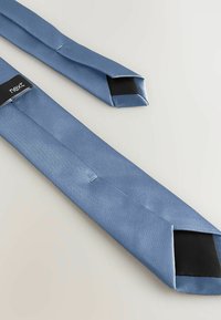 Cravate en satin bleu clair avec doublure intérieure noire et étiquette de marque « next » sur un fond clair uni.