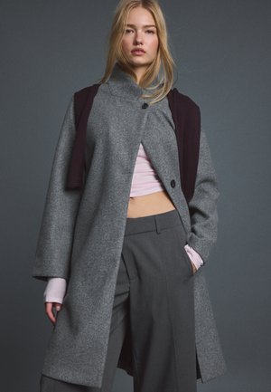 Jeune femme aux longs cheveux blonds portant un manteau gris sur un pantalon gris et un crop top rose, debout devant un fond gris uni.