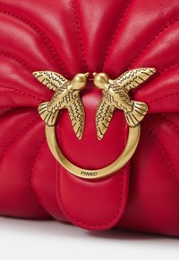 Borsa a tracolla in pelle rossa con design trapuntato, caratterizzata da due decorazioni a forma di uccello dorato sopra un anello di hardware circolare dorato.