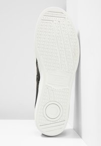 Sneaker nera con tomaia testurizzata e suola in gomma bianca, caratterizzata da un motivo a zigzag e un accento circolare. Vista dall'alto su uno sfondo uniforme.