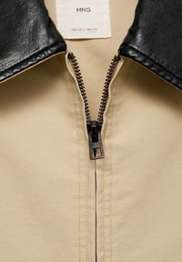 Beige Jacke mit einem schwarzen Kunstlederkragen, zentralem Reißverschluss mit bronzenen Beschlägen und sauberer Naht entlang der Seiten.