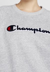 Sudadera gris hecha de tela suave, con un logo de "Champion" en negro y rojo bordado en la parte frontal, y con un cuello acanalado y mangas tipo raglán.